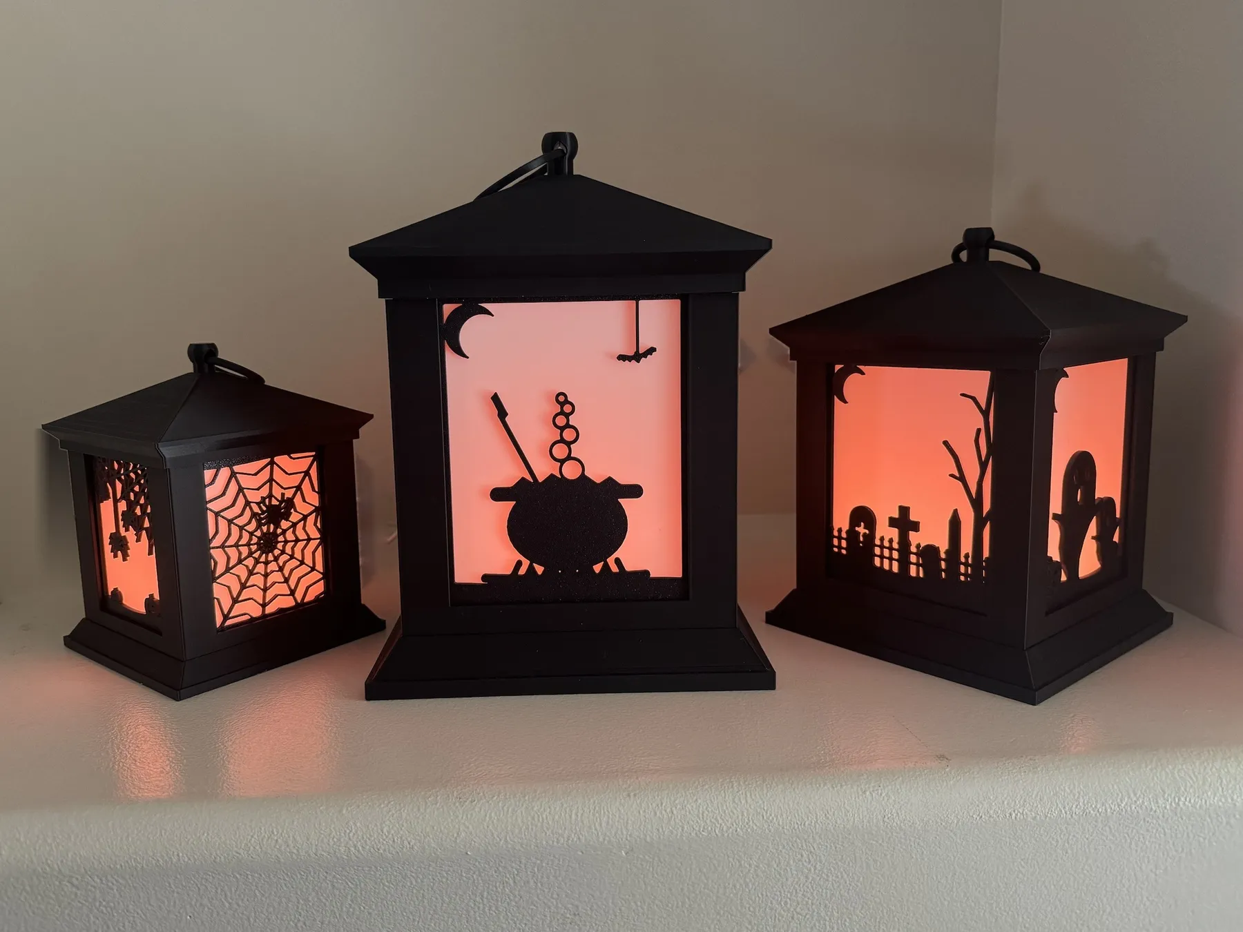 Three black lanterns glowing orange: a spider web, a witch's cauldron and a graveyard